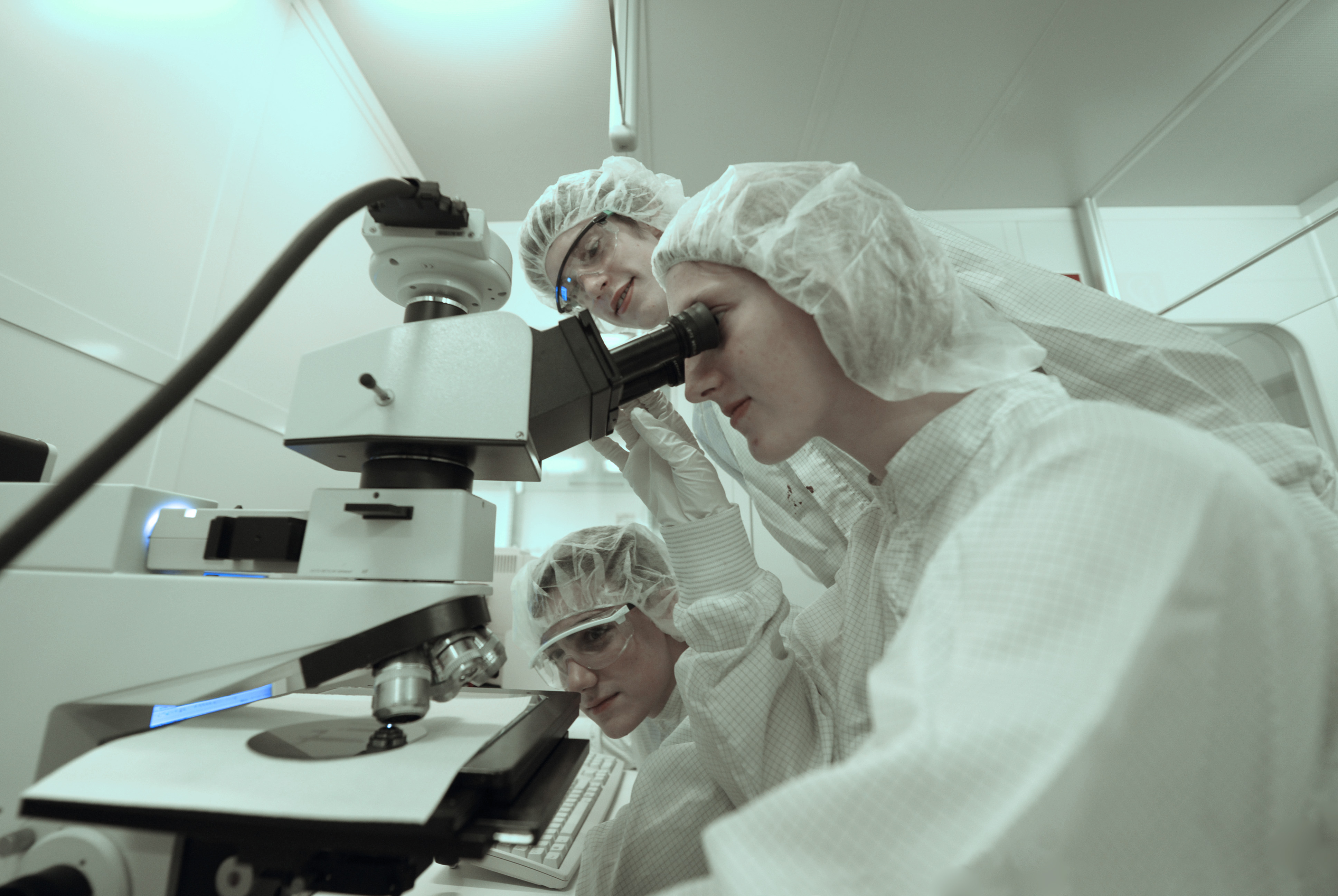Several researchers stand around a microscope in a laboratory wearing white protective suits.