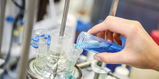 A hand fills a turquoise blue liquid into another glass vessel in the laboratory.