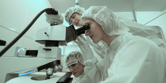 Several researchers stand around a microscope in a laboratory wearing white protective suits.