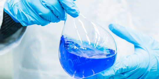 Zwei Hände in blauen Gummihandschuhen halten einen Messkolben, der mit blauer Flüssigkeit gefüllt ist.